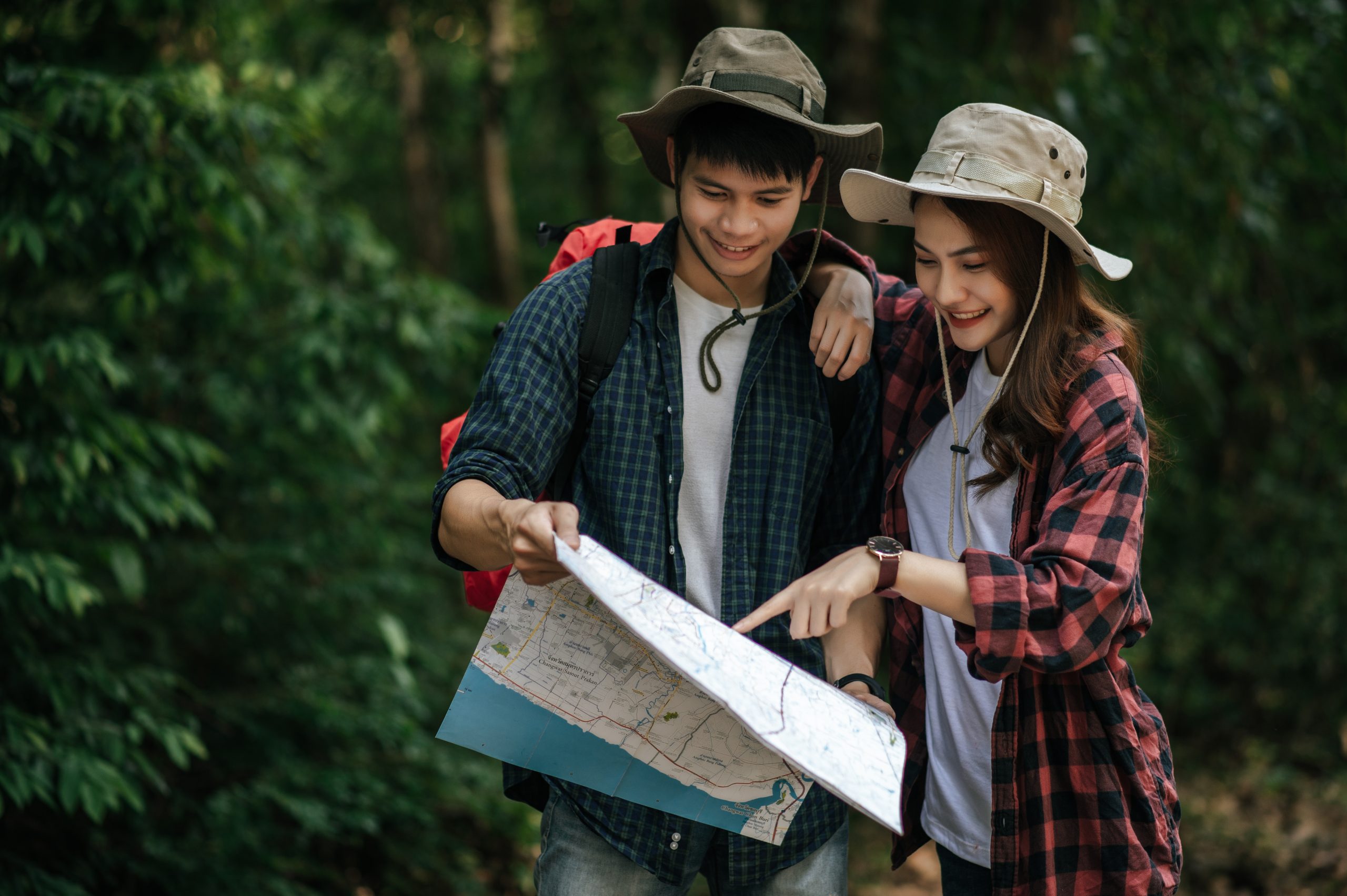 Portrait Young asian handsome man with backpack and trekking hat and pretty girlfriend standing and checking direction on paper map while walking on forest trail, backpack travel concept